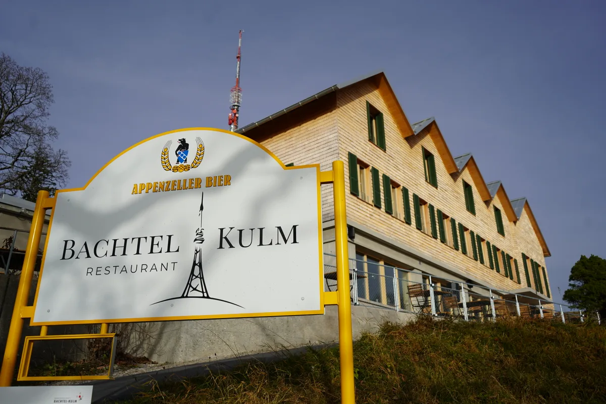 Fassade des Restaurants Bachtel-Kulm zusammen mit einer Werbetafel für das Lokal.